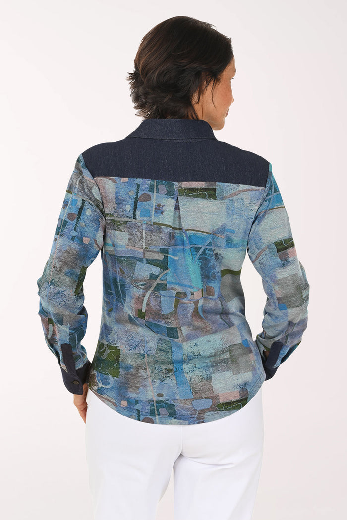 A smiling woman wearing a long-sleeve button-up shirt with an abstract blue, gray, and green patchwork print, paired with white pants, against a plain light background.