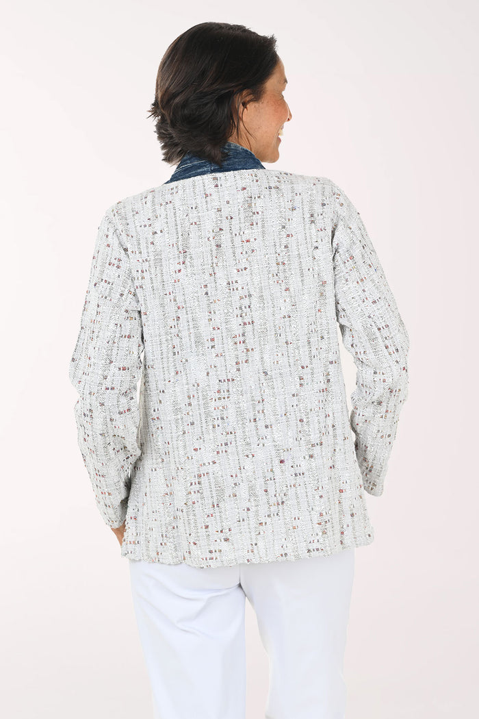 A smiling woman wearing a light gray speckled open-front cardigan with dark blue denim-style trim and front pockets, paired with a white top and white pants, against a plain light background.