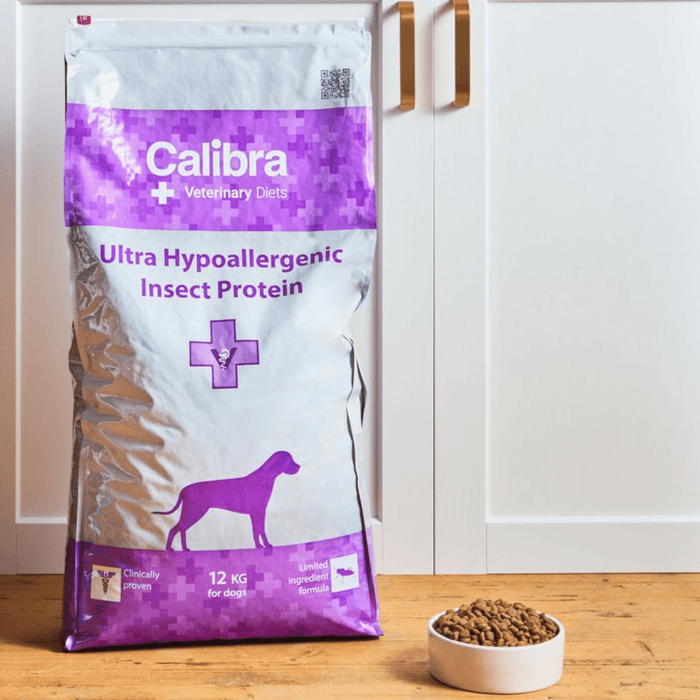 A 12 kg bag of Calibra Veterinary Diets Ultra Hypoallergenic Insect Protein dog food stands on a wooden floor next to a small white bowl filled with kibble, set against a white kitchen cabinet background.