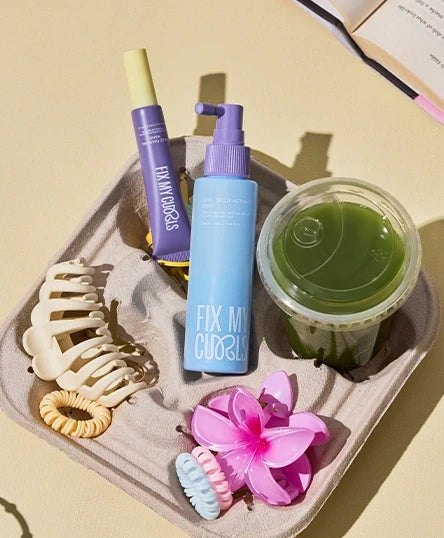 The tray contains Curl Gelly Activator Spray and Frizz Finishing Wand Duo, a green coloured drink, flower shaped hair clip and other accessories