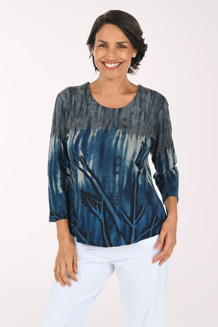 A smiling woman wearing a blue and gray tie-dye long-sleeve top with white pants, standing against a plain white studio background.