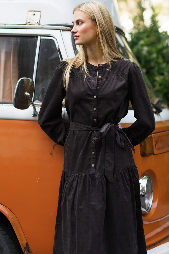 Close-up of a model wearing the Aspiga Dropped Waist Baby Cord Midi Dress in Chocolate, showing the tie belt and button placket