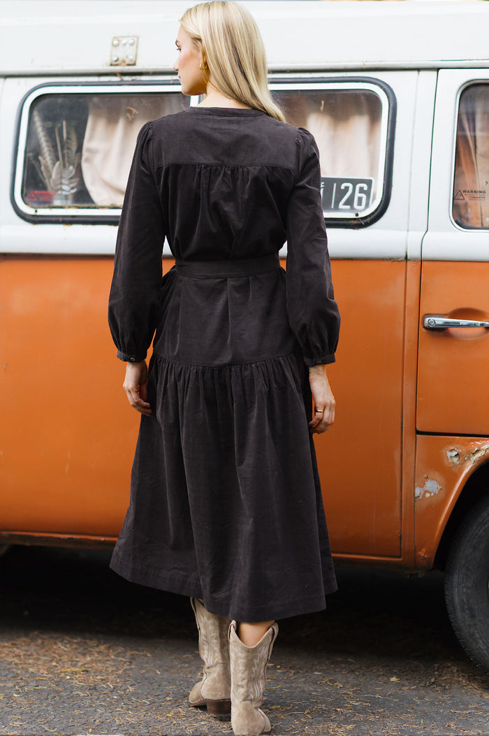 Model wearing the Aspiga Dropped Waist Baby Cord Midi Dress in Chocolate with a tie belt, back view, walking away from a vintage van