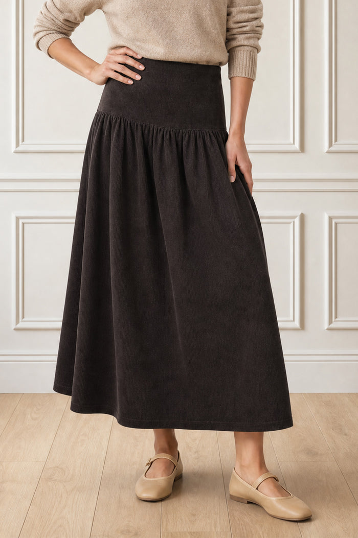 Model wearing the Aspiga Dropped Waist Baby Cord Skirt in Chocolate with a beige jumper, cropped front view, hand on hip