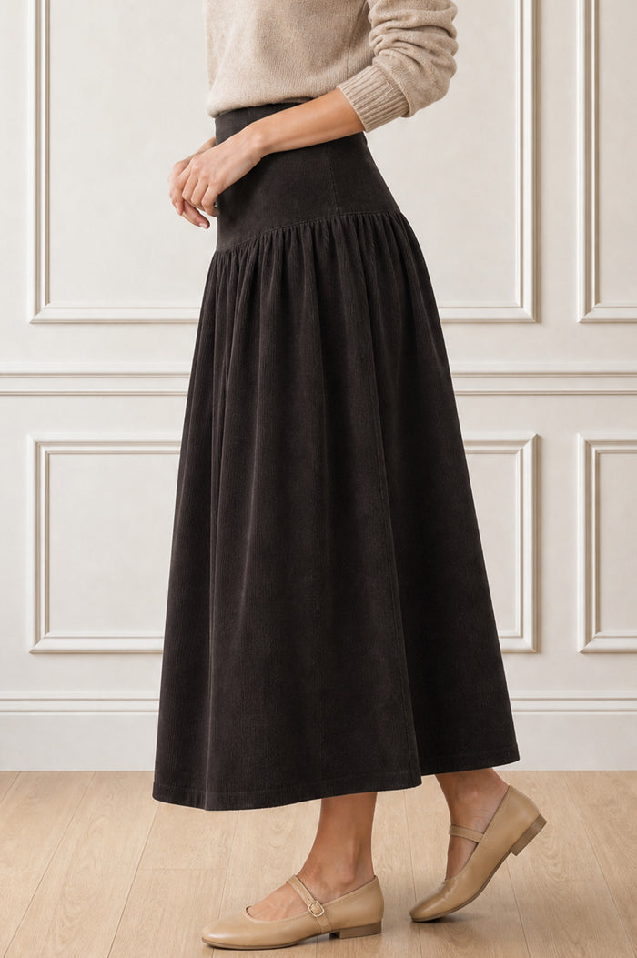 Model wearing the Aspiga Dropped Waist Baby Cord Skirt in Chocolate with a beige jumper, side view walking