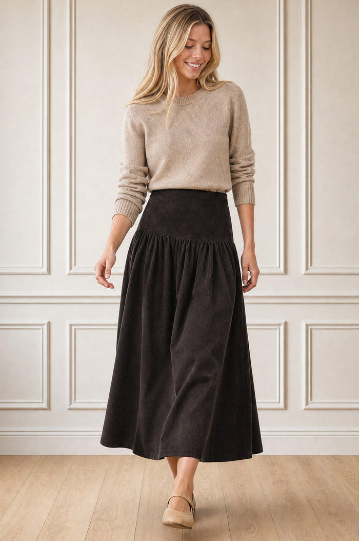Model wearing the Aspiga Dropped Waist Baby Cord Skirt in Chocolate with a beige jumper, full-length front view, smiling