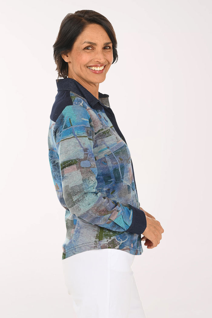 A smiling woman wearing a long-sleeve button-up shirt with an abstract blue, gray, and green patchwork print, paired with white pants, against a plain light background.