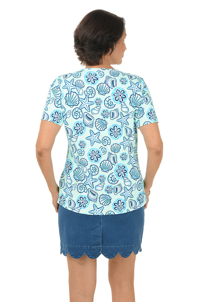 Woman wearing a blue patterned blouse with a shell design and a scallop denim skort on a white background. Back shot.