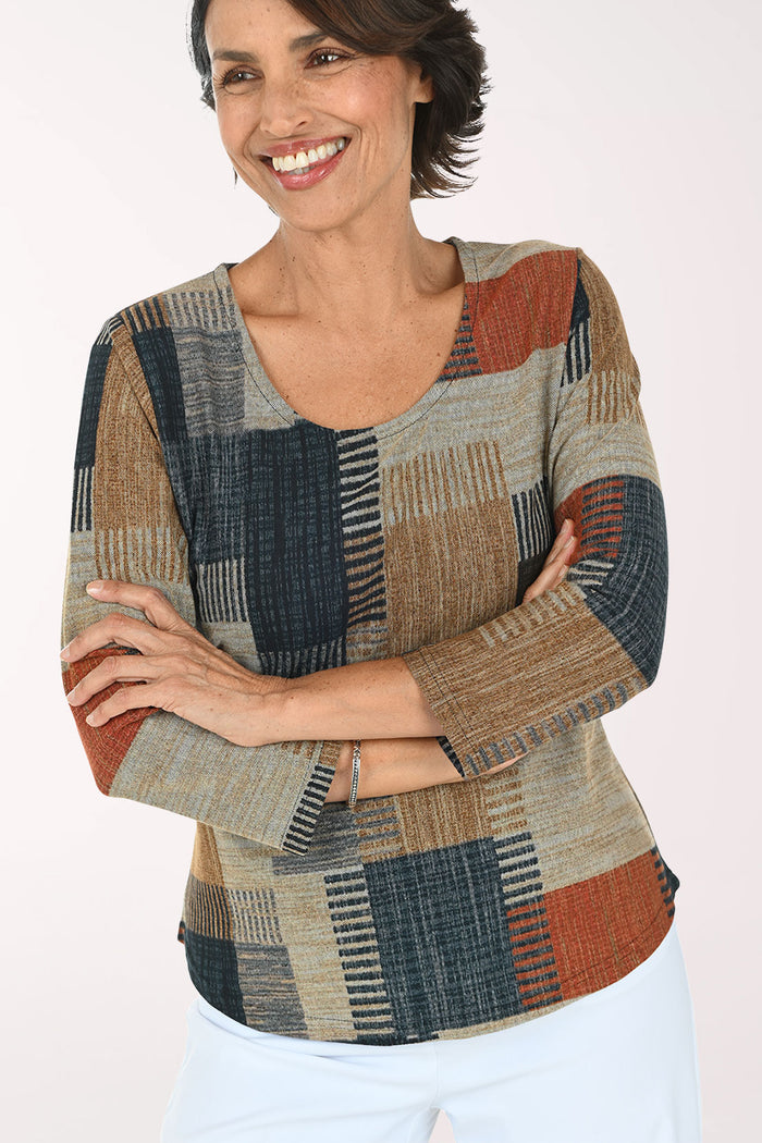 A woman modeling a three-quarter sleeve top with a patchwork geometric pattern in beige, navy, rust orange, and brown, paired with white pants against a plain white background.