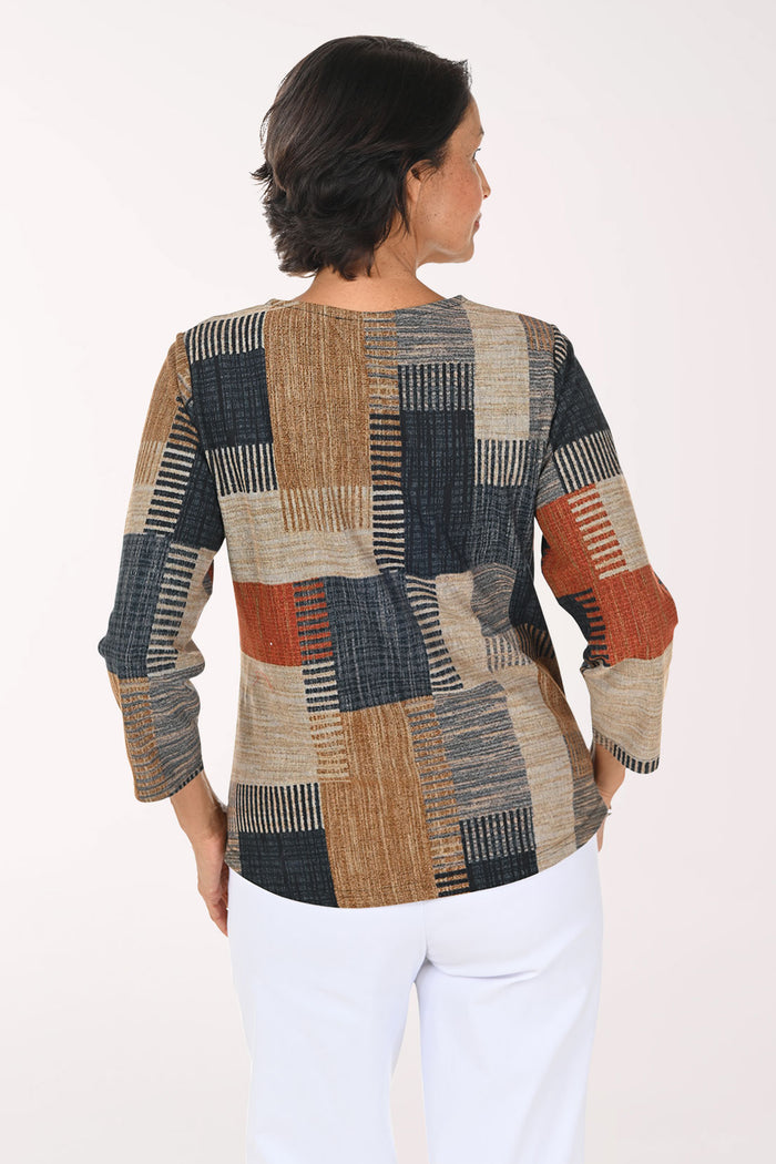 A woman modeling a three-quarter sleeve top with a patchwork geometric pattern in beige, navy, rust orange, and brown, paired with white pants against a plain white background.