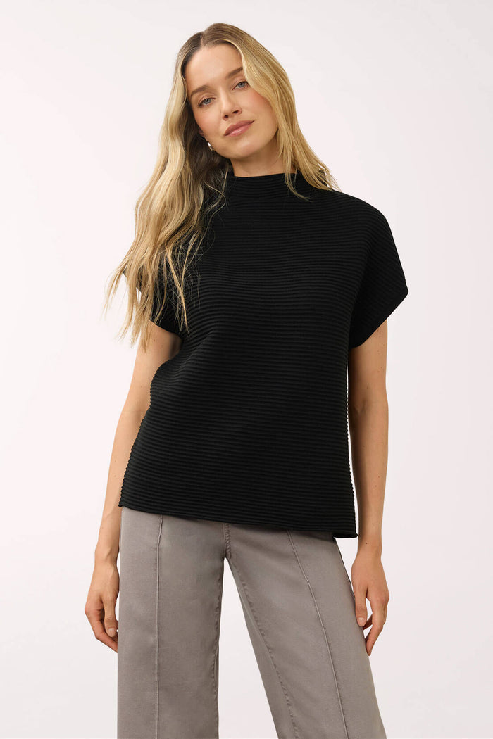 A woman modeling a black short-sleeve ribbed mock neck top with light gray pants, photographed against a plain white studio background.