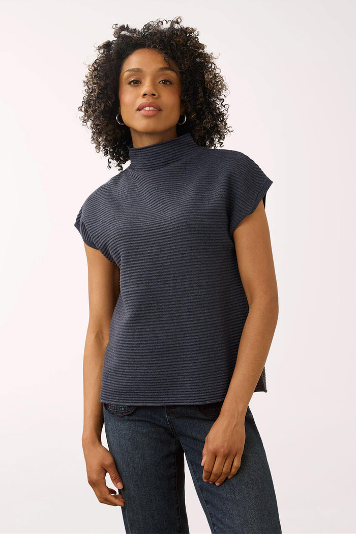 A woman with curly dark hair wearing a dark navy ribbed knit mock neck top with cap sleeves and dark blue jeans, standing against a plain light background.