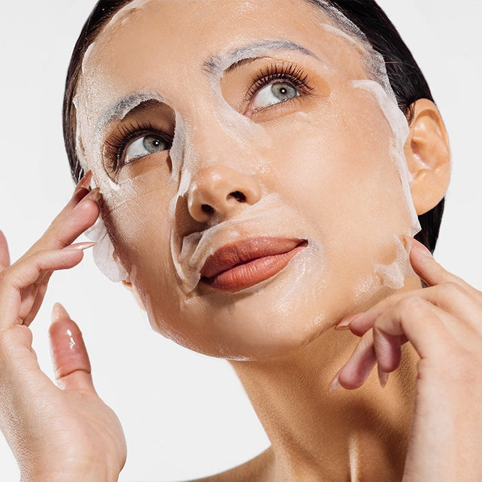 Woman gently applying a hydrating sheet mask to her face for skincare and wellness using the Restorative Facial Sheet Mask Set - 2 Mask Kit | 7e Wellness.