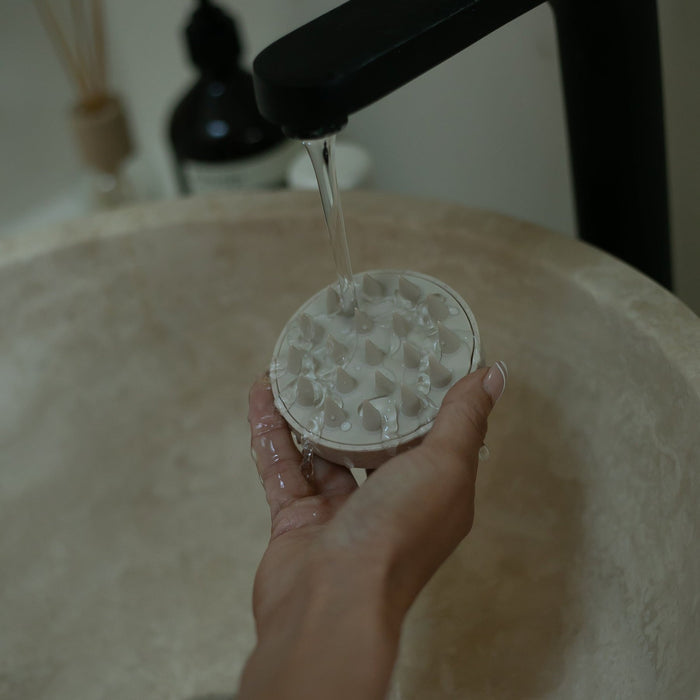 Hand holding the Restorative Scalp Massager | Vie Healing under running water above a bathroom sink, showcasing its waterproof design for easy cleaning and wellness care.
