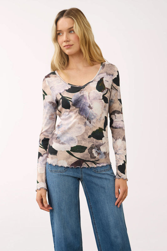 A woman modeling a long-sleeve scoop-neck top with a muted floral print in beige, gray, black, and lavender, paired with blue jeans against a plain white studio background.