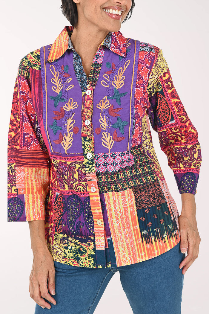 A woman modeling a vibrant, multicolored patchwork button-up shirt with intricate botanical and geometric patterns, paired with blue jeans against a plain white background.