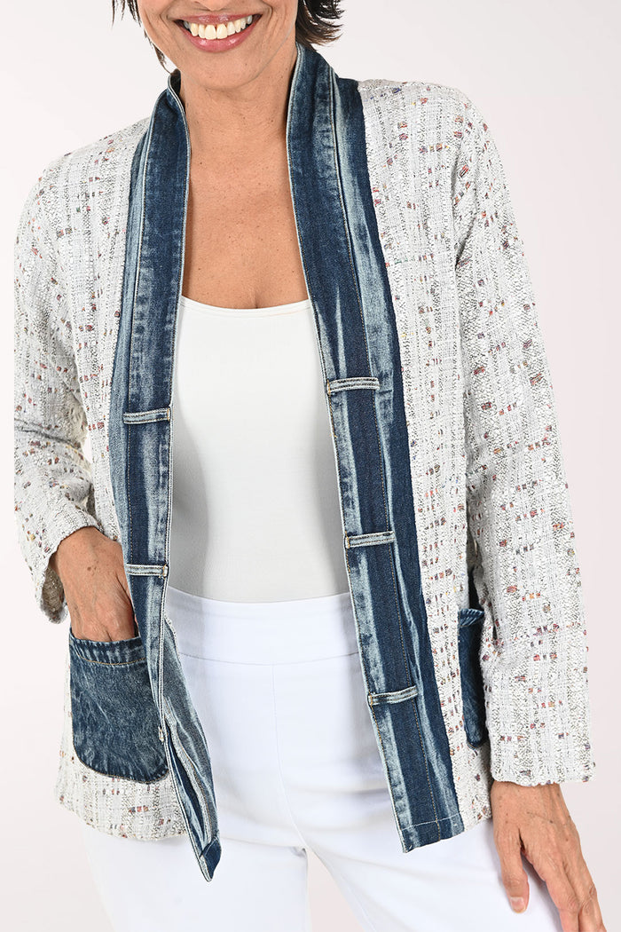 A smiling woman wearing a light gray speckled open-front cardigan with dark blue denim-style trim and front pockets, paired with a white top and white pants, against a plain light background.