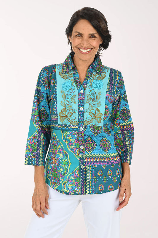 A smiling woman wearing a vibrant, multicolored patchwork print blouse with three-quarter sleeves and white pants, photographed against a solid light background.