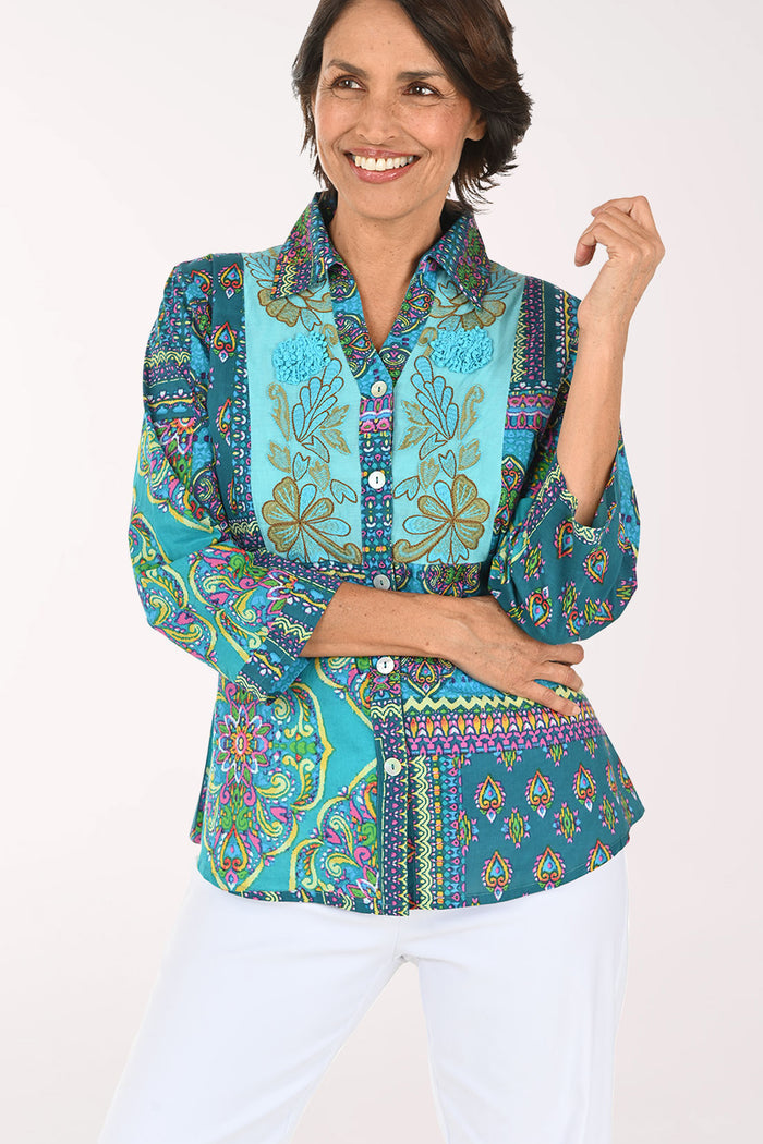 A smiling woman wearing a vibrant, multicolored patchwork print blouse with three-quarter sleeves and white pants, photographed against a solid light background.